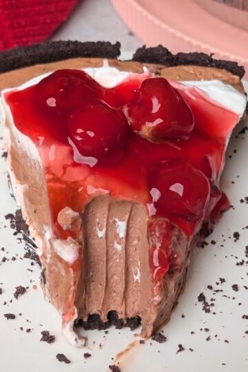 A slice of Black Forest pie with a chocolate crust, creamy chocolate filling, whipped cream, and cherry topping on a white plate with a bite taken out of it.